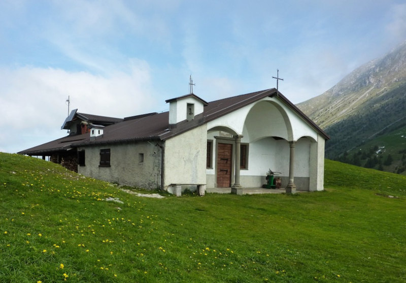 CHIESETTA DI SAN FERMO - L’oratorio di San Fermo (a cui e associato nella documentazione San Rustico, entrambi molto conosciuti nella terraferma veneta quali "magnanimi campioni della milizia cristiana") è posto a 1868 metri di altitudine, "sulle cime d’alto monte distante dal comune sette miglia in circa d’erta e scabrosa strada". Nella tradizione orale camuna, la figura di Fermo viene fatta risalire all’epoca di Carlo Magno (VIII secolo) ed è associata ai suoi santi fratelli Glisente e Cristina, vissuti rispettivamente sui monti di Berzo e di Lozio, mantenutisi tra loro in contatto mediante l’accensione di fuochi di segnalazione. Sulle tre montagne vennero edificate altrettante chiesette a loro dedicate, per garantire la possibilita di una sosta spirituale e di un ricovero materiale ai malgari e ai pastori che riempivano i pascoli durante la stagione della monticazione. È probabile che l’insediamento di San Fermo sia germinato durante la vigorosa fioritura eremitica registrata tra la ñne del secolo XI e gli inizi del XIII. All’oratorio, "fabricato ab immemorabili", la popolazione si recava "il giorno di San Lorenzo processionalmente" per cantarvi "la messa e vespri". Mantenuto in passato dalle collette offerte "spontaneamente d’anno in anno", venne completamente rifatto nel 1663 a spese del benefattore Giovan Martino Dabeni (Borno 1606 - 1693). La festa del Santo (che cade il 9 agosto) e particolarmente sentita, anche dalla comunità civica e dai villeggianti; la messa celebrata in loco nel corso della mattinata è preceduta, la sera antecedente, da una suggestiva fiaccolata che si snoda lungo i sentieri dell’alpe fino al paese, colorando di fuochi notturni l’intera montagna. (Da "La Chiesa Parrocchiale di Borno - Storia e Arte", a cura di O. Franzoni) - vedi articolo